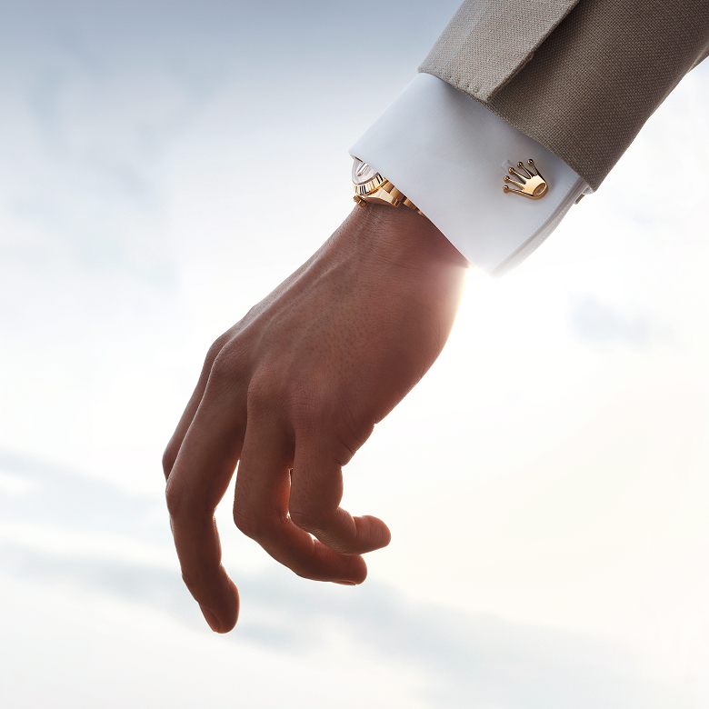 Close-up of gold Rolex Crown cufflinks worn on a shirt cuff alongside a Rolex watch.